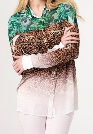 Frau mit langen blonden Haaren trägt ein Hemd mit Knöpfen, oben grüne Blumentop, in der Mitte Leopardenmuster und unten weißem, durchsichtigem Stoff, die Arme verschränkt.