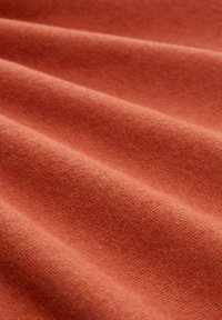 Weiches, rost-orangefarbenes Gewebe mit feiner Textur, das in sanften Falten gezeigt wird und seine glatte sowie leicht flauschige Oberfläche betont.