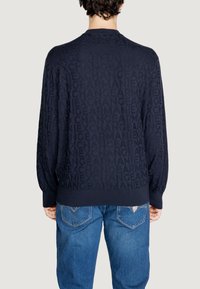 Maglione blu navy con motivo testuale tonale "ARMANI EXCHANGE", polsini e orlo a coste, e scollo rotondo. Indossato sopra jeans blu chiaro.