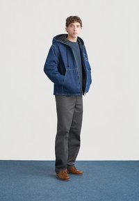 Μπουφάν από τζιν με κουκούλα, με πλαϊνές τσέπες και επένδυση με καπιτονέ, συνδυασμένο με γκρι παντελόνια και καφέ παπούτσια, που στέκεται σε μπλε χαλί.