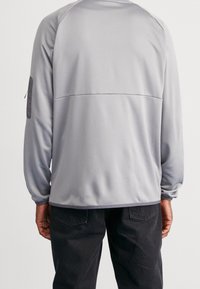 Šedý top s dlouhým rukávem se strukturovaným povrchem, obsahující horizontální šev na zádech a nášivku na levé paži.