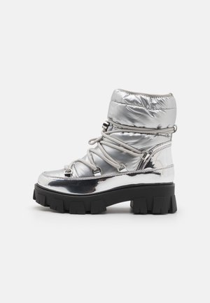 Srebrne ocieplane zimowe buty z wiązaniami obejmującymi cholewkę, masywną czarną podeszwą z bieżnikiem i wysokim kołnierzem przy kostce na białym tle.