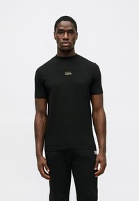 Μαύρη βαμβακερή μπλούζα με κοντά μανίκια, στρογγυλή λαιμόκοψη, με ένα μικρό χρυσό λογότυπο στο στήθος, φορεμένη με μαύρο παντελόνι.