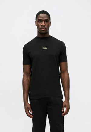 Schwarzes Baumwoll-T-Shirt mit kurzen Ärmeln, rundem Ausschnitt, mit einem kleinen, goldenen Logo auf der Brust, getragen mit schwarzen Hosen.