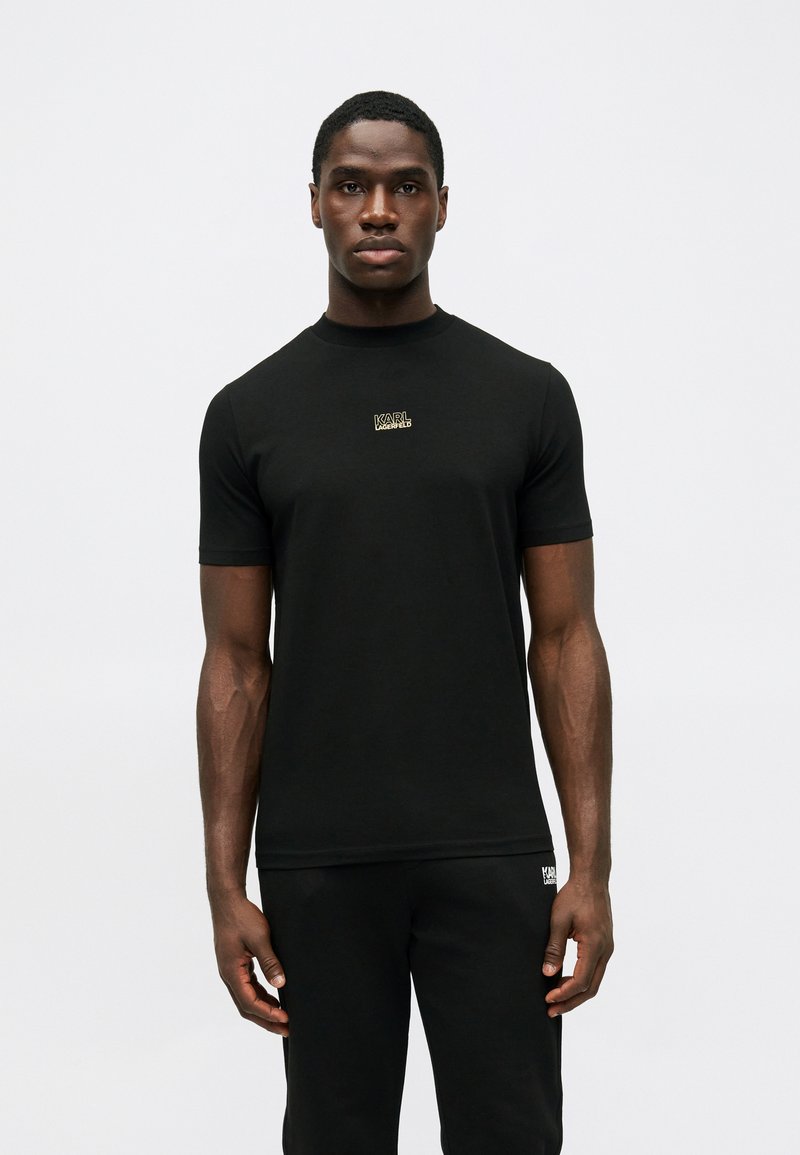 Μαύρη βαμβακερή μπλούζα με κοντά μανίκια, στρογγυλή λαιμόκοψη, με ένα μικρό χρυσό λογότυπο στο στήθος, φορεμένη με μαύρο παντελόνι.