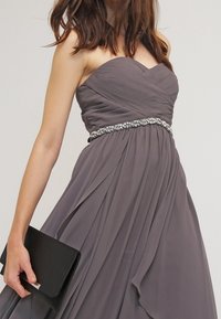 Žena v šedých plisovaných šatech bez ramínek se zdobeným pasem, drží černou kabelku v levé ruce.