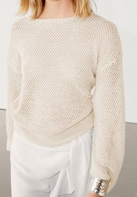 Lysebeige, åbenstrikket sweater med lange ærmer og bred halsudskæring; har en skinnende sølvmanchet ved ærmeenderne.