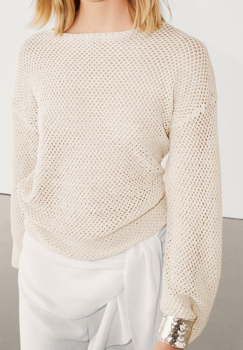 Lysebeige, åbenstrikket sweater med lange ærmer og bred halsudskæring; har en skinnende sølvmanchet ved ærmeenderne.