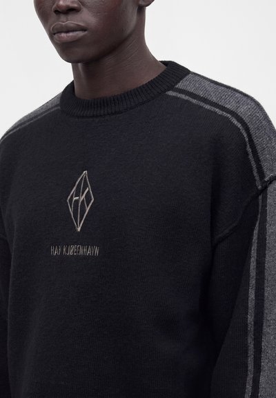 Gros plan d’une personne portant un pull noir avec des rayures grises sur les manches et un logo géométrique avec du texte sur la poitrine.