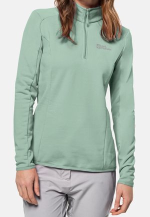 Vrouw draagt een lichtgroene fleece trui met lange mouwen en ritssluiting en lichtgrijze broek, staand tegen een witte achtergrond.