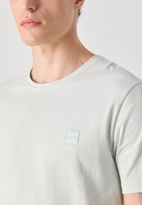 T-shirt verde-claro de gola redonda feito de algodão suave, apresentando um pequeno patch de logotipo quadrado no peito esquerdo. Textura suave, mangas curtas.