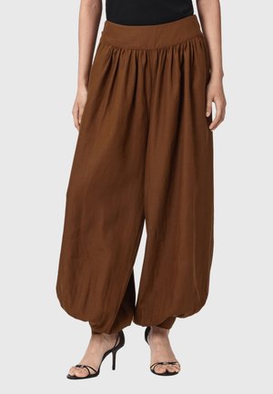 Vrouw draagt losse, bruine, wijde broek met hoge taille, een geplooide tailleband en zwarte sandalen met hoge hakken.