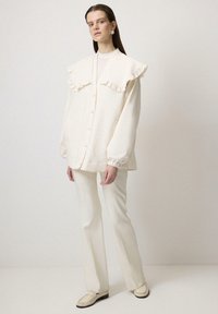 Camisa blanca de botones con cuello rizado y mangas largas, combinada con pantalones blancos a juego y zapatos de color claro, sobre un fondo neutro.
