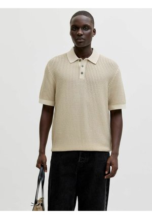 Bărbat purtând un tricou polo bej texturat cu mâneci scurte și pantaloni negri, ținând o geantă bej cu mânere negre pe un fundal simplu.