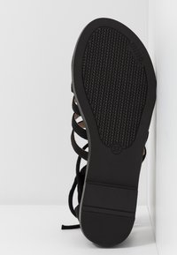 Sandale noire avec une semelle texturée, dotée de brides croisées et d'une étiquette de taille (37). Surface supérieure lisse avec un minimum de détails.