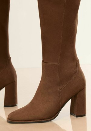 Botte en suède marron montée jusqu'au genou avec un bout carré et un talon épais. Présente un design sans couture et des détails de couture subtils.