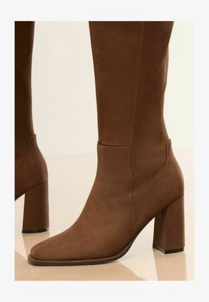 Pruun sametiste säärebootsid ruudukujulise varbaga ja massiivse kontsaga. Omab sujuvat disaini ja peeneid õmbluse detaile.