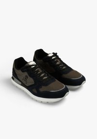 Pareja de zapatillas negras y verde olivo con suelas blancas y un logo de calavera y tibias cruzadas en el costado, colocadas sobre un fondo blanco.