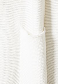 Vitt stickat tröjtyg med ett strukturerat mönster. Har en rullad kant och en synlig söm, som visar ett mjukt, medeltungt material.