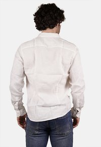 Camicia bianca in lino con maniche lunghe, polsini allacciati e una vestibilità leggermente rilassata. La schiena presenta un dettaglio a yoke e una superficie liscia.