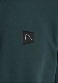 Mörkgrön sweatshirt med en slät textur, med en liten svart fyrkantig lapp med en vit logotypdesign på framsidan.