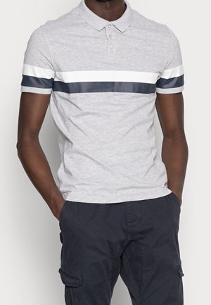Mann trägt ein hellgraues Polo-Shirt mit weißen und dunkelblauen horizontalen Streifen und dunkelblaue Cargohosen, die Hände in den Taschen.