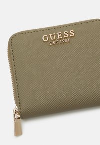 Grønn syntetisk lommebok med preget tekstur, gull "Guess"-logo, glidelåslukking og en dekorativ glidelåstrekk.