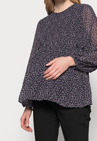 Blusa nera floreale in tessuto leggero e trasparente; presenta motivi rosa e bianchi sparsi, maniche lunghe con polsini arricciati e una vestibilità comoda.