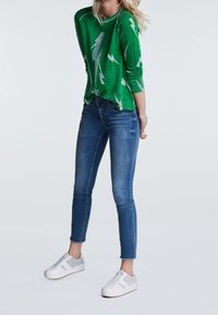 Pull vert avec un motif d'éclair blanc, associé à un jean bleu ajusté et des baskets blanches avec des accents gris. Tenue décontractée présentée.
