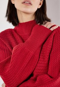 Chandail en tricot rouge avec un col haut à côtes et un motif texturé. Le tissu semble doux avec un design douillet et épais.