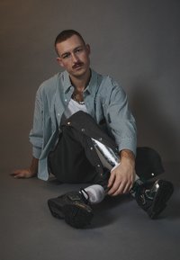 Csíkos zöld és fehér gombos ing, szürke nadrág fényes ezüst részletekkel, fekete sportcipő, ülő póz egy szürke háttér előtt.