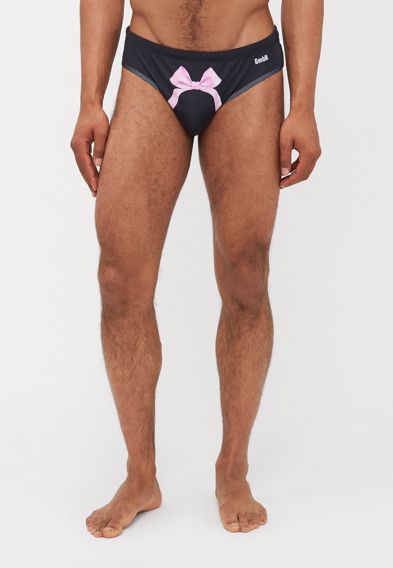 Bikini noir avec un détail de nœud rose à l'avant, dotée d'une coupe ajustée et d'une texture lisse. Le nom de la marque est imprimé sur le côté.