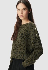 Vrouw met lang bruin haar, gekleed in een olijfgroene trui met een luipaardprint en zilveren knopen op de schouder, en zwarte broek.