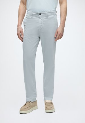 Homme portant un pantalon slim gris clair et des chaussures en daim beige, debout devant un fond blanc uni.