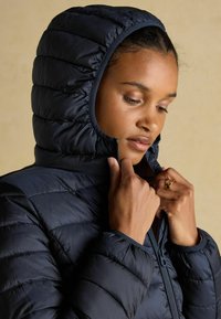 Sinine puhvis jope, millel on tikande muster ja sobiv kapuuts. Ees on lukuga sulgemine, sile kangas ja kontrastsed detailid.