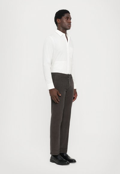Uomo in piedi di profilo che indossa una camicia bianca con bottoni, pantaloni marrone scuro e mocassini in pelle nera su uno sfondo chiaro e semplice.