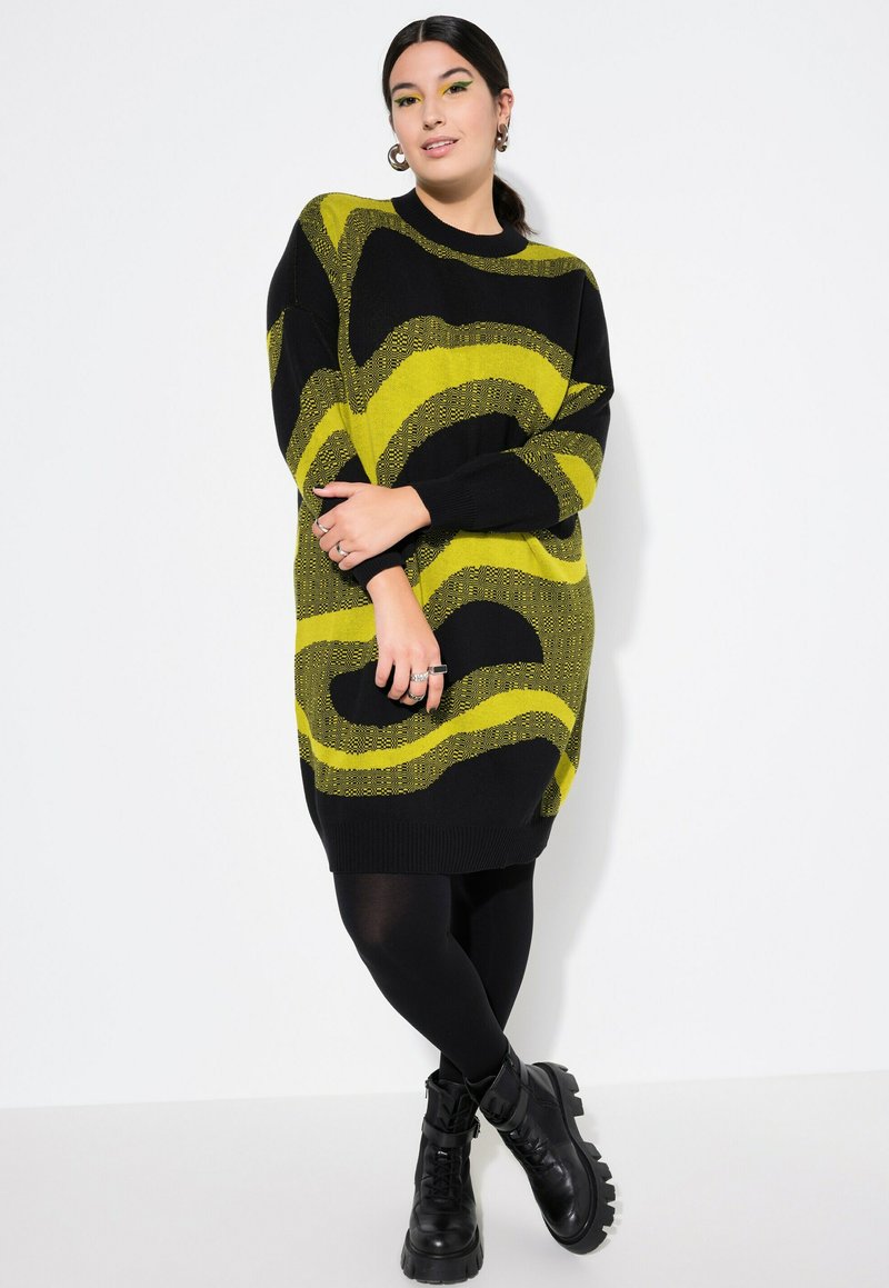 Abito maglione nero con audace motivo a onde gialle. Maniche lunghe, vestibilità oversize, rifinito con polsini e fondo a coste. Indossato sopra collant neri.