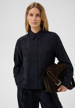 Femme aux cheveux blonds portant une chemise noire boutonnée et un pantalon noir, tenant un grand sac en daim brun foncé avec une boucle.