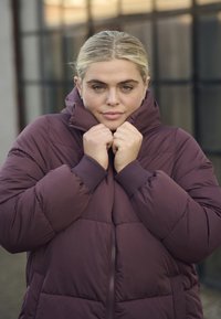 Bordo puffrová bunda s vysokým golierom, so zúženými manžetami a zipsami, zobrazená s claspom na golieri, zdôrazňujúca teplo a útulnosť.