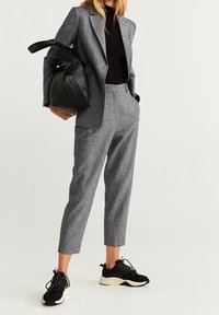 Grijze op maat gemaakte blazer over een zwart geribd topje, gecombineerd met grijze taps toelopende broek en zwarte sneakers. Een zwarte tas hangt over de arm.