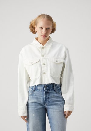 En ung person med lyst, krøllet hår har på seg en hvit, kortermet knappet jakke og høytlivte blå jeans, stående foran en ensfarget bakgrunn.