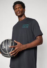 T-shirt preto de mangas curtas com uma impressão gráfica em verde-água no peito; textura suave; usado por uma pessoa segurando uma bola de basquete preta.