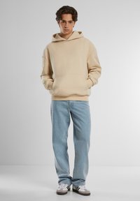 Beige hooded sweatshirt with a front pocket, paired with light blue jeans and gray sneakers. Smooth fabric texture, relaxed fit.