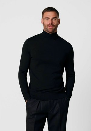 Man die een zwarte coltrui en zwarte broek draagt, poseert met zijn handen in zijn zakken tegen een effen lichte achtergrond.