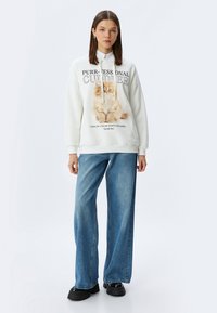 Witte sweatshirt met een grafisch ontwerp van een kat en tekst. Combinatie met lichtblauwe wijde jeans en zwarte platform schoenen. Soepele stof, casual pasvorm.
