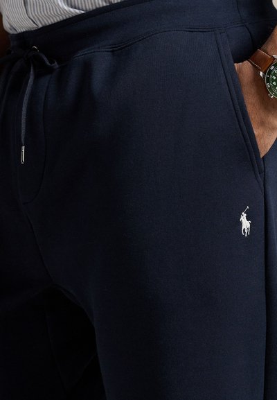 Navy blue sweatpants with elastic waistband and drawstring, featuring a white logo on the upper thigh and side pockets for convenience.