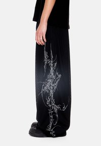 Άτομο που φοράει φαρδιά μαύρα παντελόνια με λευκό αφηρημένο σχέδιο γραμμών, στέκεται με το αριστερό χέρι χαλαρό στο πλάι και μαύρα αθλητικ ά παπούτσια σε λευκό φόντο.