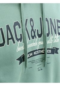 Πράσινο φούτερ με κουκούλα με τη φράση «JACK & JONES» σε μεγάλα μαύρα γράμματα και επιπλέον λευκό και μαύρο κείμενο που παρουσιάζει το σχεδιασμό και πόλεις από κάτω.