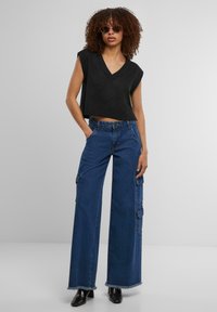 Top corto senza maniche in maglia nera con scollo a V, abbinato a pantaloni cargo in denim a gamba larga con tasche laterali e orli sfilacciati.