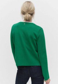 Grön långärmad topp i mjukt material, med rund halsringning och en liten metallaccent på baksidan. Rakt snitt, avslappnad design.
